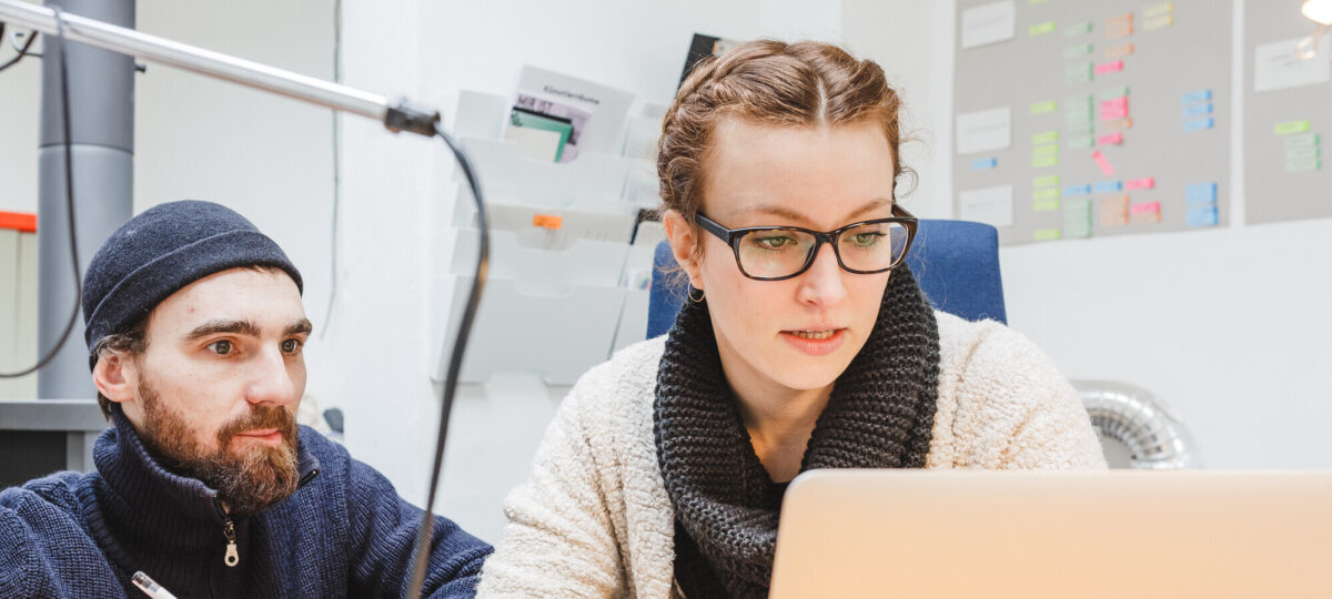 Zwei Personen arbeiten gemeinsam an einem Schreibtisch. Eine Frau mit Brille und Wollschal schaut konzentriert auf einen Laptop, während ein Mann mit Mütze und Bart neben ihr sitzt, einen Stift in der Hand hält und aufmerksam zuhört. Im Hintergrund sind eine Wand mit bunten Post-its und Ablagefächer zu sehen.