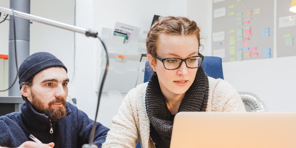 Gründung Zwei Personen arbeiten gemeinsam an einem Schreibtisch. Eine Frau mit Brille und Wollschal schaut konzentriert auf einen Laptop, während ein Mann mit Mütze und Bart neben ihr sitzt, einen Stift in der Hand hält und aufmerksam zuhört. Im Hintergrund sind eine Wand mit bunten Post-its und Ablagefächer zu sehen.