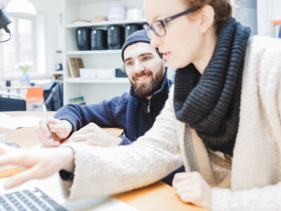 Zwei Personen arbeiten gemeinsam an einem Schreibtisch. Im Vordergrund zeigt eine Frau mit Brille und Wollschal auf den Laptopbildschirm, während ein lächelnder Mann mit Mütze und Bart daneben sitzt und Notizen macht. Im Hintergrund sind Regale und ein Fenster sichtbar.
