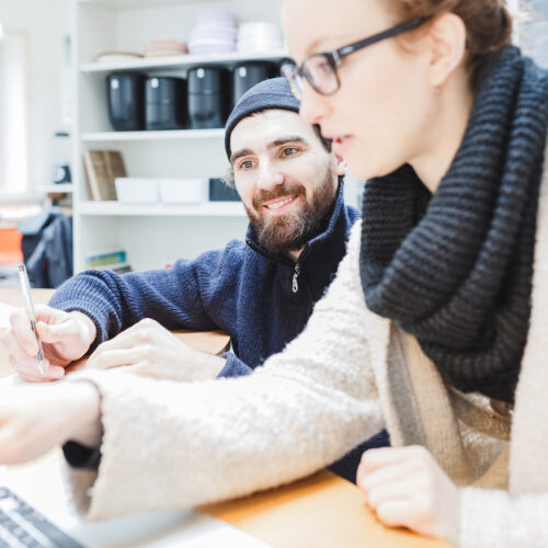 Zwei Personen arbeiten gemeinsam an einem Schreibtisch. Im Vordergrund zeigt eine Frau mit Brille und Wollschal auf den Laptopbildschirm, während ein lächelnder Mann mit Mütze und Bart daneben sitzt und Notizen macht. Im Hintergrund sind Regale und ein Fenster sichtbar.