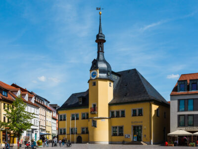 Historisches Rathausgebäude in Apolda mit markantem gelbem Anstrich und einem zentralen Uhrturm mit geschwungener Spitze und Wetterfahne. Das Gebäude ist umgeben von bunt gestrichenen Wohn- und Geschäftshäusern sowie modernen Gebäuden. Auf dem Platz davor spazieren Menschen bei sonnigem Wetter, einige sitzen im Außenbereich eines Cafés. Der Himmel ist klar und blau.