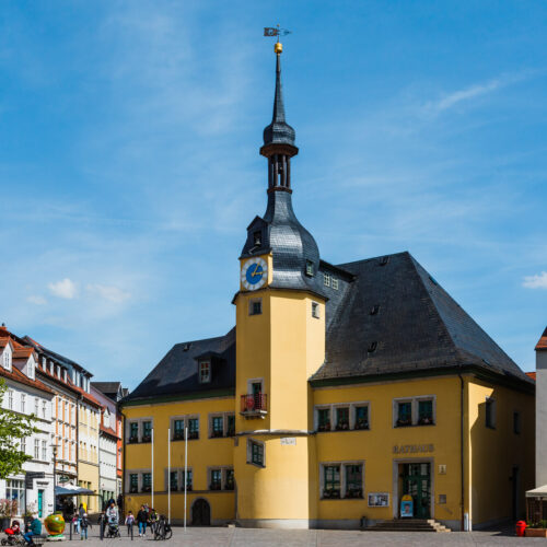 Historisches Rathausgebäude in Apolda mit markantem gelbem Anstrich und einem zentralen Uhrturm mit geschwungener Spitze und Wetterfahne. Das Gebäude ist umgeben von bunt gestrichenen Wohn- und Geschäftshäusern sowie modernen Gebäuden. Auf dem Platz davor spazieren Menschen bei sonnigem Wetter, einige sitzen im Außenbereich eines Cafés. Der Himmel ist klar und blau.