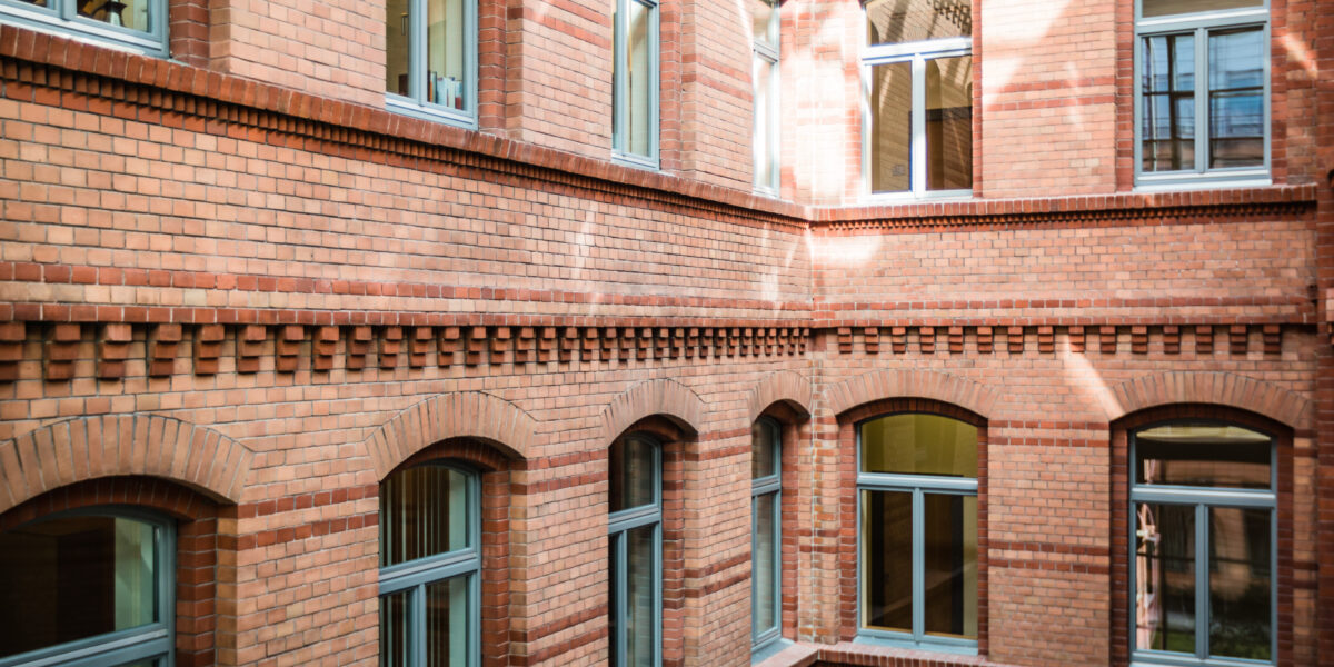 TAB-Gebäude innen Innenhof eines historischen Backsteingebäudes mit mehreren Fenstern. Die Fassade besteht aus roten Ziegelsteinen mit dekorativen Backsteinmustern und Rundbogenfenstern. Sonnenlicht fällt durch ein Glasdach oder eine Lichtöffnung und erzeugt helle Reflexionen auf der Wand.