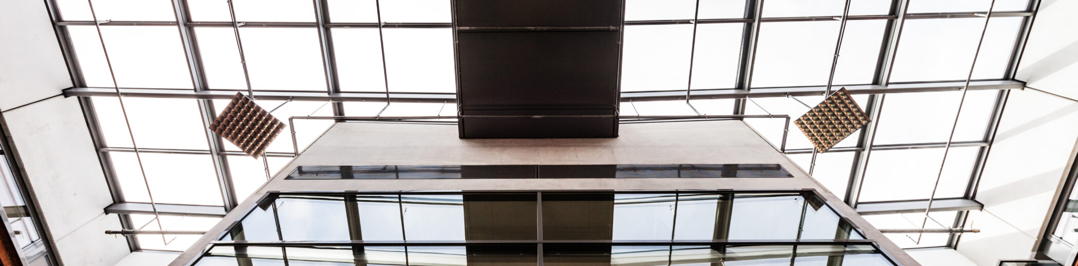 Blick von unten in ein modernes Atrium mit Glasfassade und Glasdach. In der Mitte ragt ein schmaler, rechteckiger Brückengang über die Gebäudeseiten. Reflektionen in den Fenstern zeigen die Struktur des Daches. Mehrere quadratische Akustik- oder Lichtpaneele hängen von der Decke.