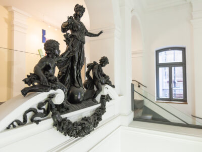 TAB-Gebäude Figuren Innenansicht eines historischen Gebäudes mit weiß gestrichenen Wänden. Im Vordergrund befindet sich eine detailreiche dunkle Skulpturengruppe auf einem Treppenabsatz. Die Skulptur zeigt eine stehende Frauengestalt auf einer Kugel, flankiert von zwei kindlichen Figuren. Die Brüstung ist mit floralen Verzierungen und einem Kranz aus dunklem Material geschmückt. Im Hintergrund ist ein Fenster mit grauem Rahmen sowie ein Treppenaufgang mit Glasgeländer zu sehen.