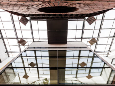 TAB-Gebäude Blick von unten in ein modernes Atrium mit Glasfassade und Glasdach. In der Mitte ragt ein schmaler, rechteckiger Brückengang über die Gebäudeseiten. Reflektionen in den Fenstern zeigen die Struktur des Daches. Mehrere quadratische Akustik- oder Lichtpaneele hängen von der Decke.