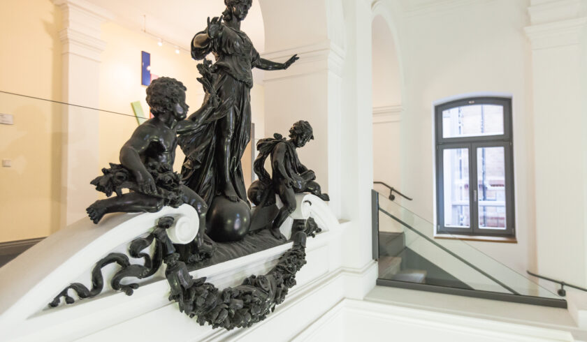 TAB-Gebäude Figuren Innenansicht eines historischen Gebäudes mit weiß gestrichenen Wänden. Im Vordergrund befindet sich eine detailreiche dunkle Skulpturengruppe auf einem Treppenabsatz. Die Skulptur zeigt eine stehende Frauengestalt auf einer Kugel, flankiert von zwei kindlichen Figuren. Die Brüstung ist mit floralen Verzierungen und einem Kranz aus dunklem Material geschmückt. Im Hintergrund ist ein Fenster mit grauem Rahmen sowie ein Treppenaufgang mit Glasgeländer zu sehen.