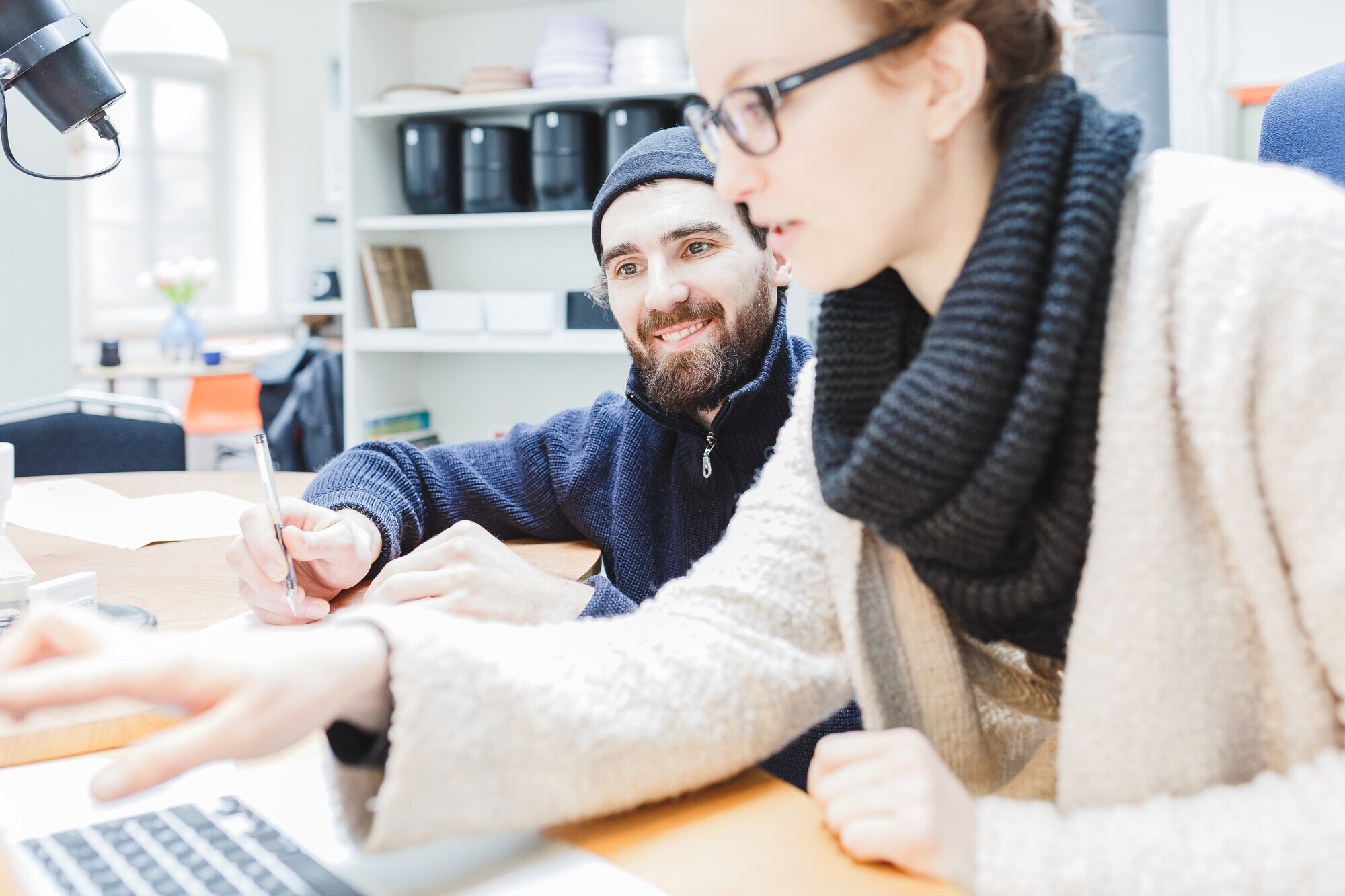 Zwei Personen arbeiten gemeinsam an einem Schreibtisch. Im Vordergrund zeigt eine Frau mit Brille und Wollschal auf den Laptopbildschirm, während ein lächelnder Mann mit Mütze und Bart daneben sitzt und Notizen macht. Im Hintergrund sind Regale und ein Fenster sichtbar.