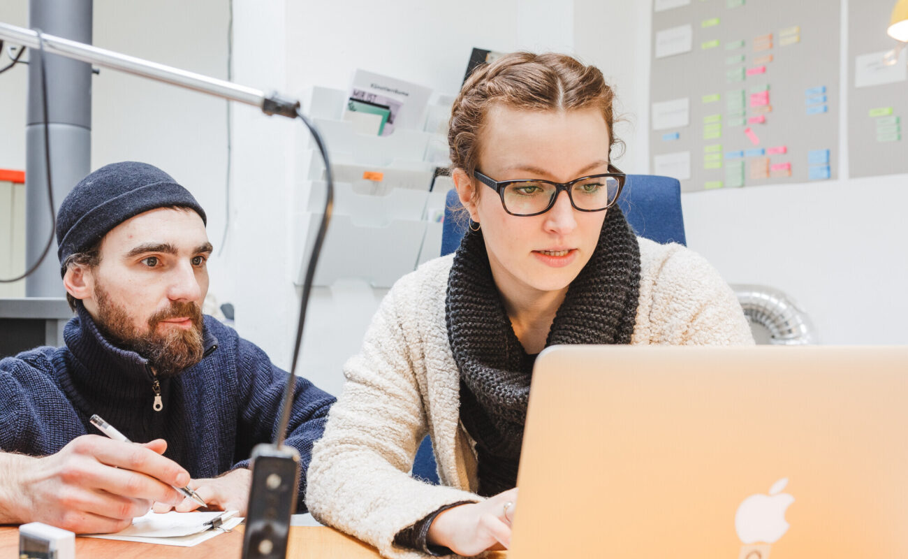 Zwei Personen arbeiten gemeinsam an einem Schreibtisch. Eine Frau mit Brille und Wollschal schaut konzentriert auf einen Laptop, während ein Mann mit Mütze und Bart neben ihr sitzt, einen Stift in der Hand hält und aufmerksam zuhört. Im Hintergrund sind eine Wand mit bunten Post-its und Ablagefächer zu sehen.