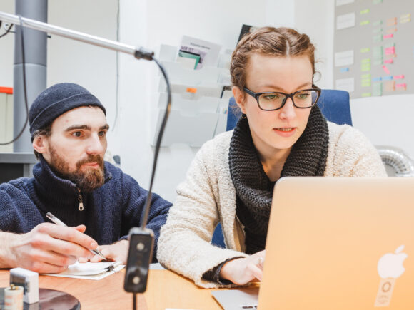 Zwei Personen arbeiten gemeinsam an einem Schreibtisch. Eine Frau mit Brille und Wollschal schaut konzentriert auf einen Laptop, während ein Mann mit Mütze und Bart neben ihr sitzt, einen Stift in der Hand hält und aufmerksam zuhört. Im Hintergrund sind eine Wand mit bunten Post-its und Ablagefächer zu sehen.