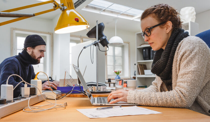 Zwei Personen arbeiten konzentriert an Laptops in einem modernen, hellen Büro. Im Vordergrund sitzt eine Frau mit Brille, Wollschal und flauschiger Jacke, im Hintergrund ein Mann mit dunklem Pullover und Mütze. Auf dem Holztisch liegen Dokumente, Kabel und eine gelbe Schreibtischlampe leuchtet den Arbeitsbereich aus.