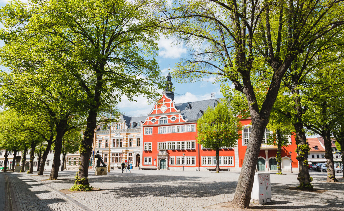 Marktplatz in Arnstadt mit dem markanten rot-weißen Rathausgebäude im Stil der Renaissance. Das Rathaus steht im Zentrum des Bildes, eingerahmt von frischen, grün belaubten Bäumen und flankiert von historischen Bürgerhäusern. Der Platz ist mit Kopfsteinpflaster bedeckt, einige Passanten spazieren oder verweilen im Schatten der Bäume. Der Himmel ist leicht bewölkt, aber hell und freundlich.