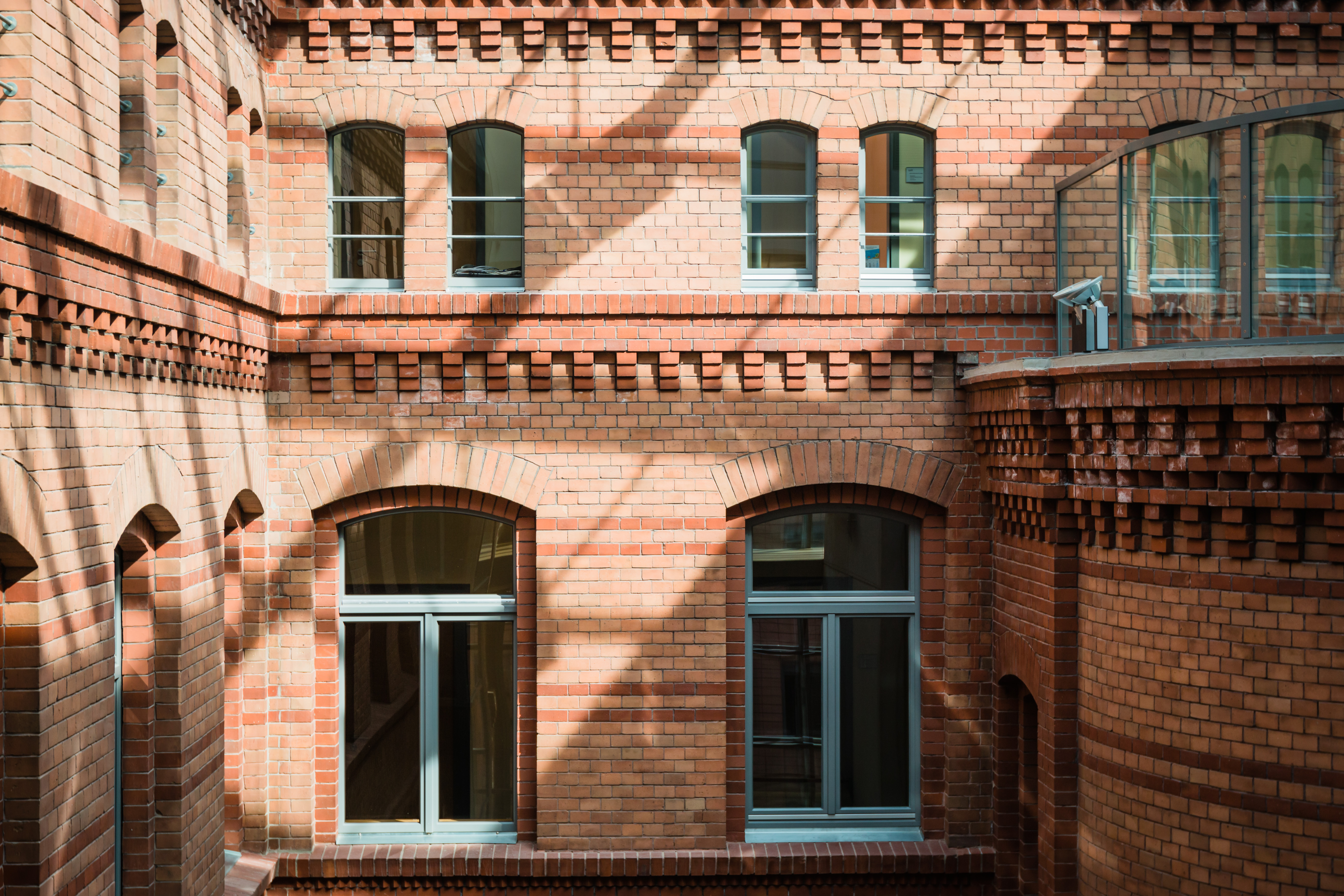 Ansicht eines Innenhofs mit historischer Backsteinfassade in warmen Rottönen. Die Fassade ist mit dekorativen Ziegelmustern verziert, darunter Zahnschnittfriese und Rundbogenfenster mit grauen Fensterrahmen. Die Glasüberdachung des Hofes wirft ein Schattenmuster auf die Wände. Rechts im Bild ist ein abgerundeter Gebäudeteil mit einer modernen Glasbrüstung zu sehen.