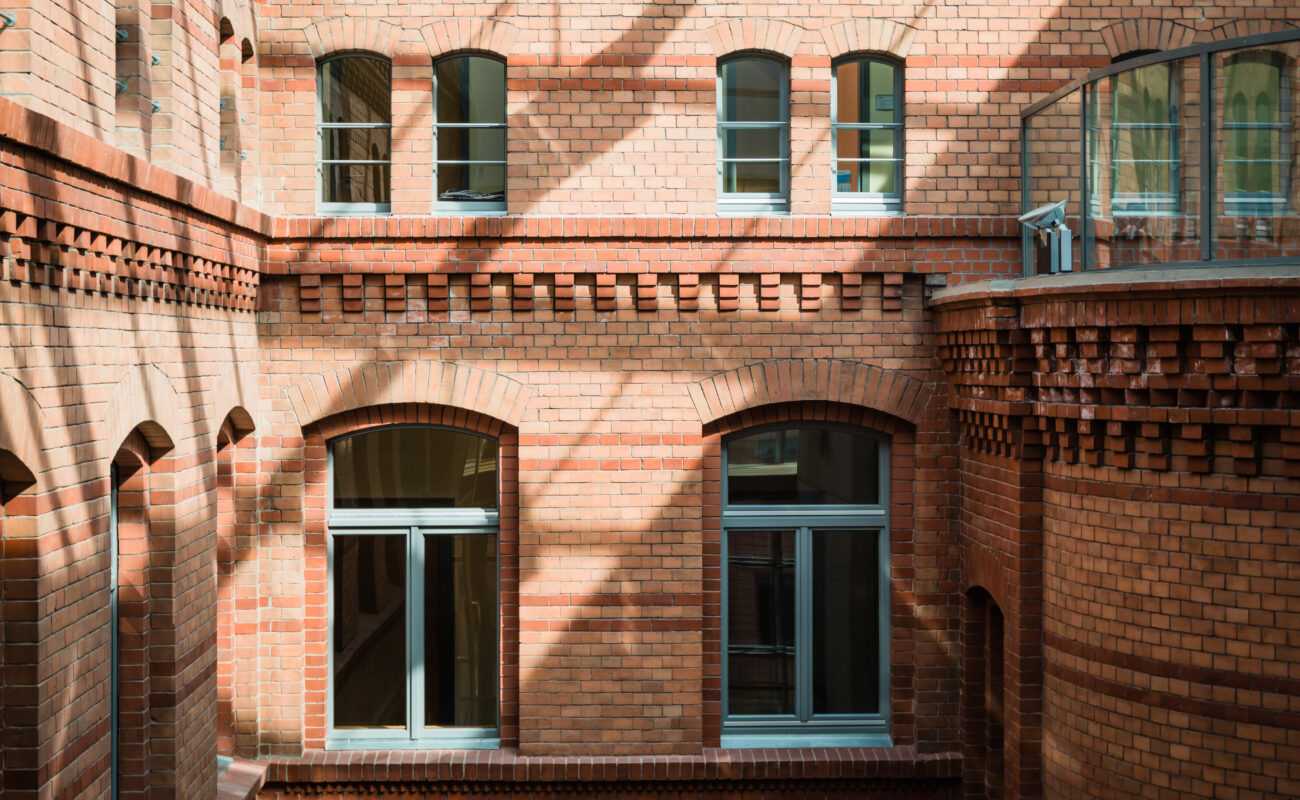 Ansicht eines Innenhofs mit historischer Backsteinfassade in warmen Rottönen. Die Fassade ist mit dekorativen Ziegelmustern verziert, darunter Zahnschnittfriese und Rundbogenfenster mit grauen Fensterrahmen. Die Glasüberdachung des Hofes wirft ein Schattenmuster auf die Wände. Rechts im Bild ist ein abgerundeter Gebäudeteil mit einer modernen Glasbrüstung zu sehen.