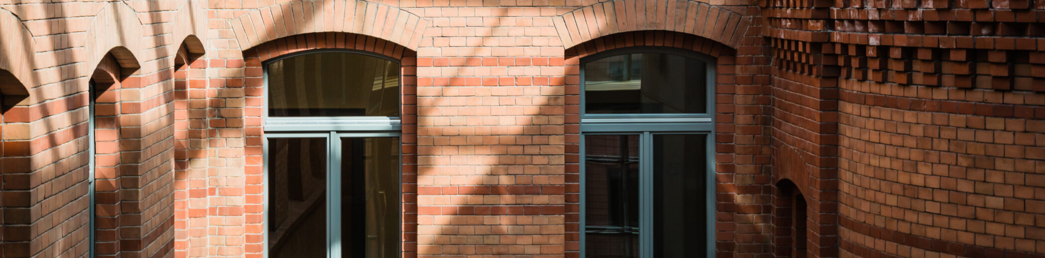 Ansicht eines Innenhofs mit historischer Backsteinfassade in warmen Rottönen. Die Fassade ist mit dekorativen Ziegelmustern verziert, darunter Zahnschnittfriese und Rundbogenfenster mit grauen Fensterrahmen. Die Glasüberdachung des Hofes wirft ein Schattenmuster auf die Wände. Rechts im Bild ist ein abgerundeter Gebäudeteil mit einer modernen Glasbrüstung zu sehen.