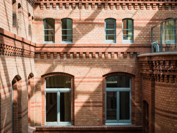 Ansicht eines Innenhofs mit historischer Backsteinfassade in warmen Rottönen. Die Fassade ist mit dekorativen Ziegelmustern verziert, darunter Zahnschnittfriese und Rundbogenfenster mit grauen Fensterrahmen. Die Glasüberdachung des Hofes wirft ein Schattenmuster auf die Wände. Rechts im Bild ist ein abgerundeter Gebäudeteil mit einer modernen Glasbrüstung zu sehen.