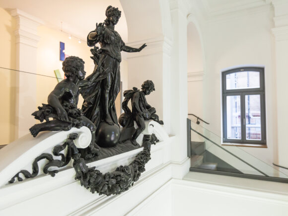 Innenansicht eines historischen Gebäudes mit weiß gestrichenen Wänden. Im Vordergrund befindet sich eine detailreiche dunkle Skulpturengruppe auf einem Treppenabsatz. Die Skulptur zeigt eine stehende Frauengestalt auf einer Kugel, flankiert von zwei kindlichen Figuren. Die Brüstung ist mit floralen Verzierungen und einem Kranz aus dunklem Material geschmückt. Im Hintergrund ist ein Fenster mit grauem Rahmen sowie ein Treppenaufgang mit Glasgeländer zu sehen.