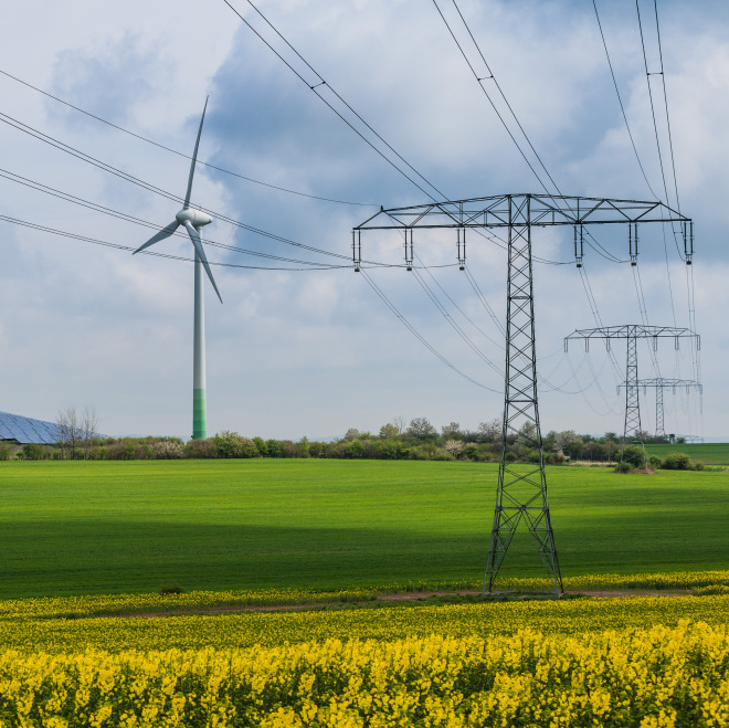 Auf dem Bild: Ein Windrad, eine Stromtrasse sowie Solarpanele auf einem Feld.