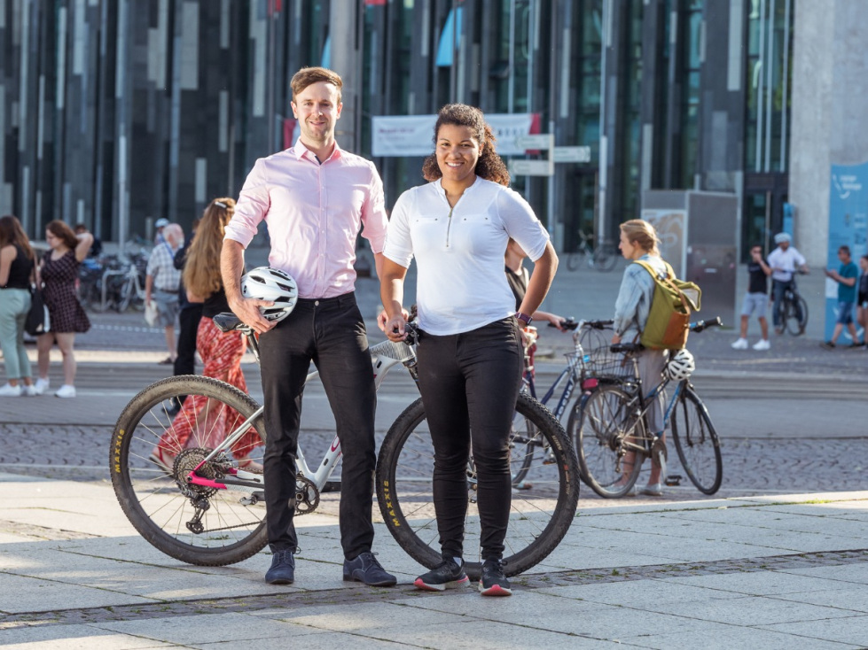 Gründungsteam Dashfactory: Lelia König und Sandro Beck (im Bild: das Team auf einem belebten Platz vor einem Fahrrad stehend)